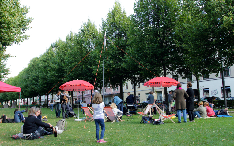Lawn on Bordeauxplatz, with a radio antenna on a mast in the middle. Grouped around it are tables with radio equipment, parasols with the word "Radio" on them and deckchairs. People sit in the chairs, stand and sit on blankets.