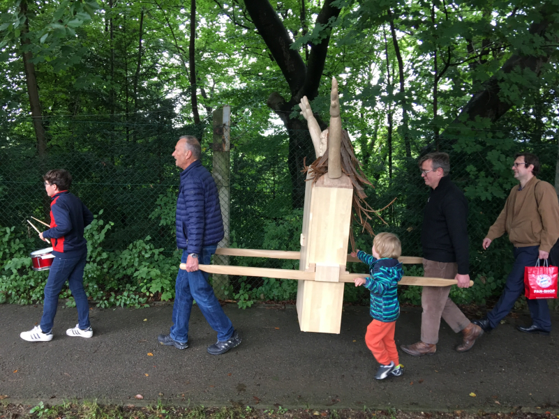 Prozession mit mehreren Menschen auf einem Spazierweg am Wald. Zwei Teilnehmer tragen als Prozessionsgegenstand eine kastenartige Frauenfigur aus Holz, die sogenannte "Konsum-Werte-Madonna".