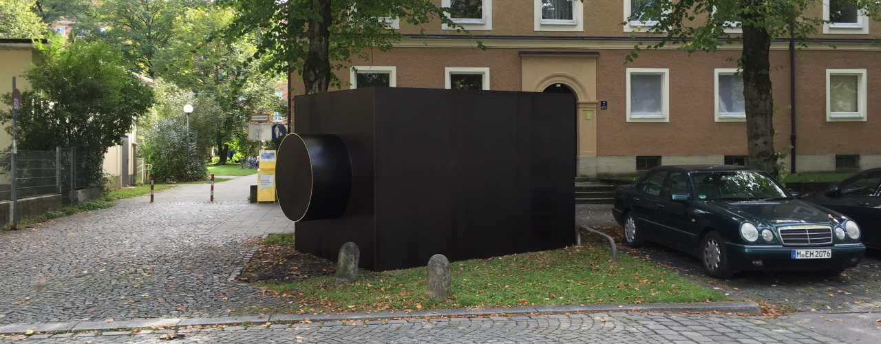 Auf einem Grünstreifen neben parkenden Autos ist eine Skulptur in Form eines dunklen, rechteckigen Baukörpers zu sehen, der an ein Kameragehäuse erinnert.