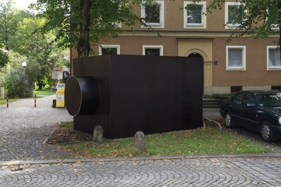 Auf einem Grünstreifen neben parkenden Autos ist eine Skulptur in Form eines dunklen, rechteckigen Baukörpers zu sehen, der an ein Kameragehäuse erinnert.