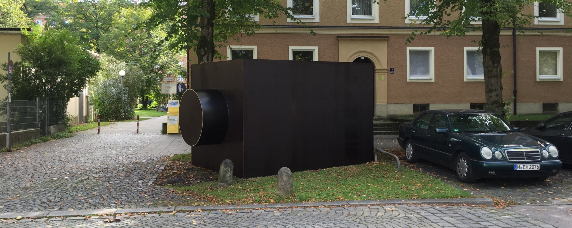 A sculpture in the form of a dark rectangular structure, reminiscent of a camera housing, can be seen on a grass verge next to parked cars.