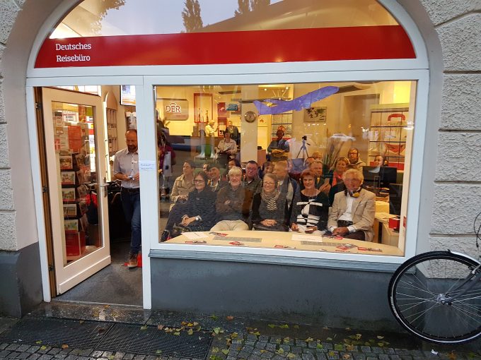 Eine Gruppe von Menschen sitzt in mehreren Stuhlreihen im Reisebüro an der Münchner Freiheit und blickt aus dem Schaufenster nach außen.