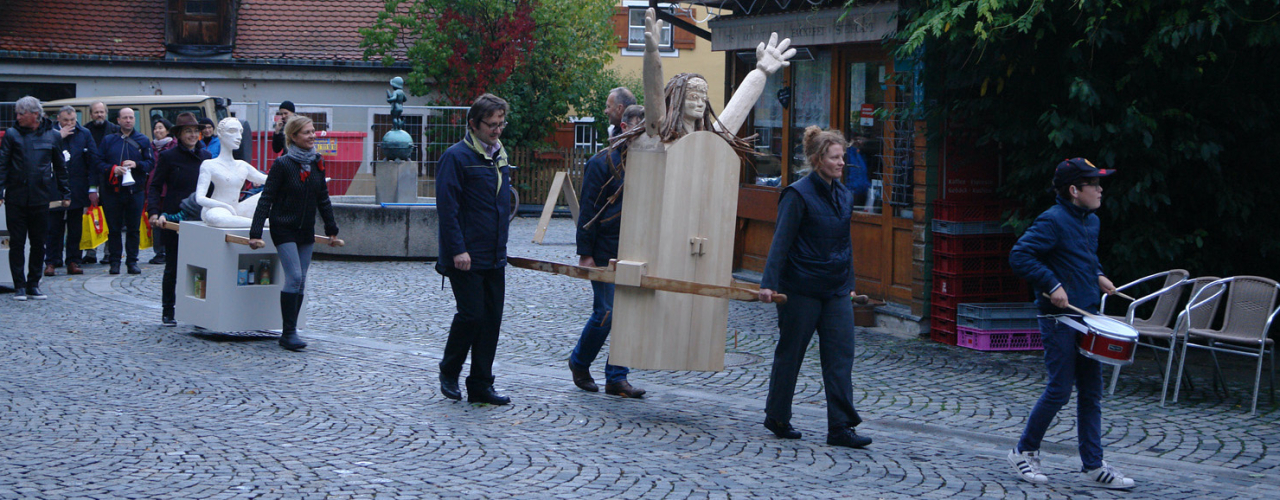 Prozession mit mehreren Menschen am Wiener Platz. Einige Teilnehmende tragen als Prozessionsgegenstände zwei Frauenfiguren, eine aus Holz und eine aus Gips, die sogenannten "Konsum-Werte-Madonnen".