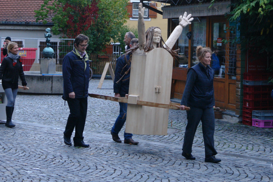 Prozession mit mehreren Menschen am Wiener Platz. Einige Teilnehmende tragen als Prozessionsgegenstände zwei Frauenfiguren, eine aus Holz und eine aus Gips, die sogenannten "Konsum-Werte-Madonnen".