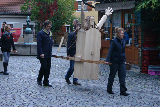 Procession with several people at Wiener Platz. Some participants carry two female figures as procession objects, one made of wood and one made of plaster, the so-called "Consumer Values Madonnas".