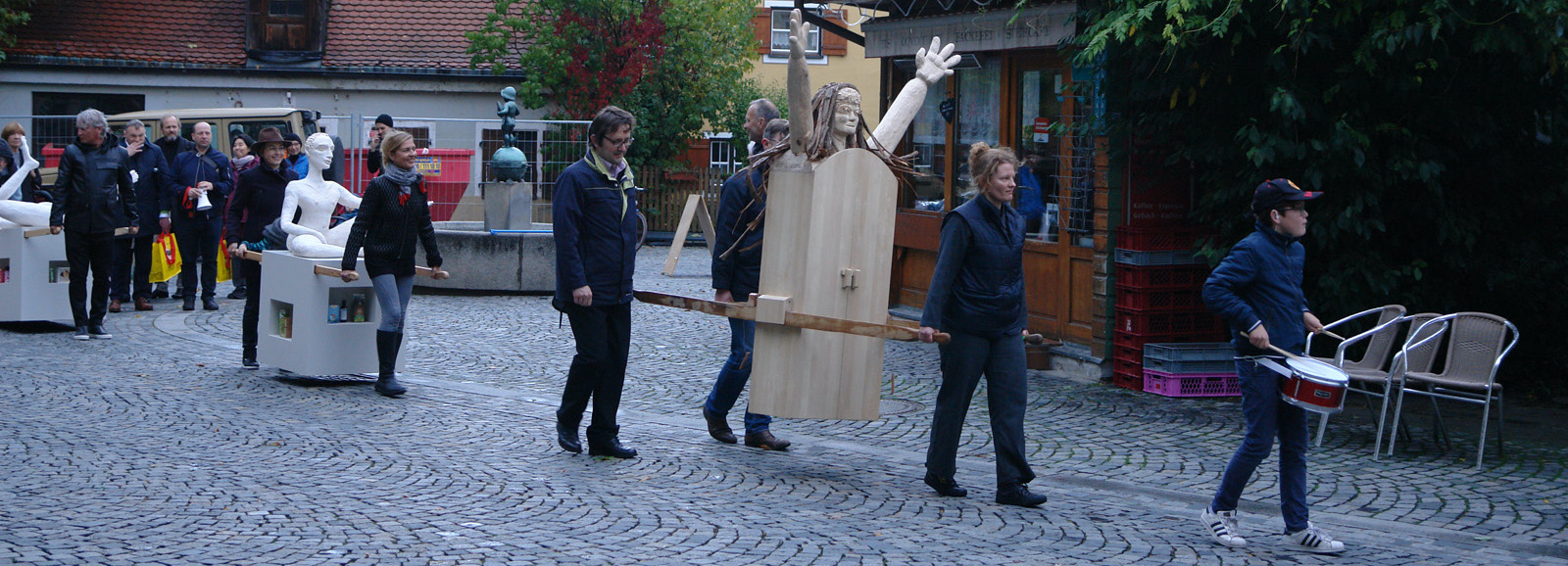 Procession with several people at Wiener Platz. Some participants carry two female figures as procession objects, one made of wood and one made of plaster, the so-called "Consumer Values Madonnas".