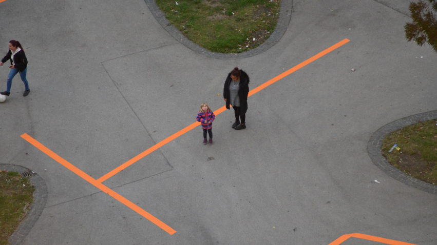 Ein orangenes "T" als Großbuchstabe ist auf den Boden geklebt. Daneben stehen eine Frau und ein Kind.