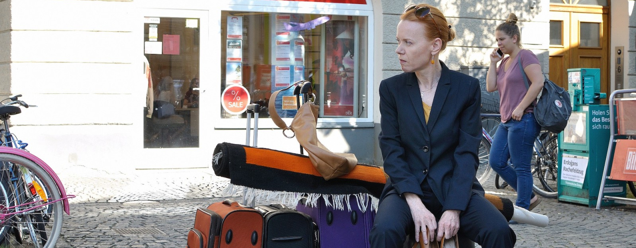 A woman in a blue suit sits on several packed suitcases and a rolled carpet in front of the travel agency at Münchner Freiheit.