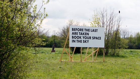 Weiße Reklametafel mit einem Holzgestell auf einer Wiese mit Bäumen. In schwarzer Schrift in Großbuchstaben zu lesen: "Before the last are taken, book your space in the sky".