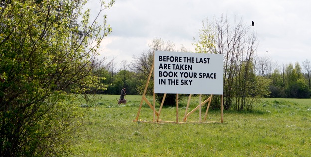 White billboard with a wooden frame on a meadow with trees. In black letters in capital letters: "Before the last are taken, book your space in the sky".