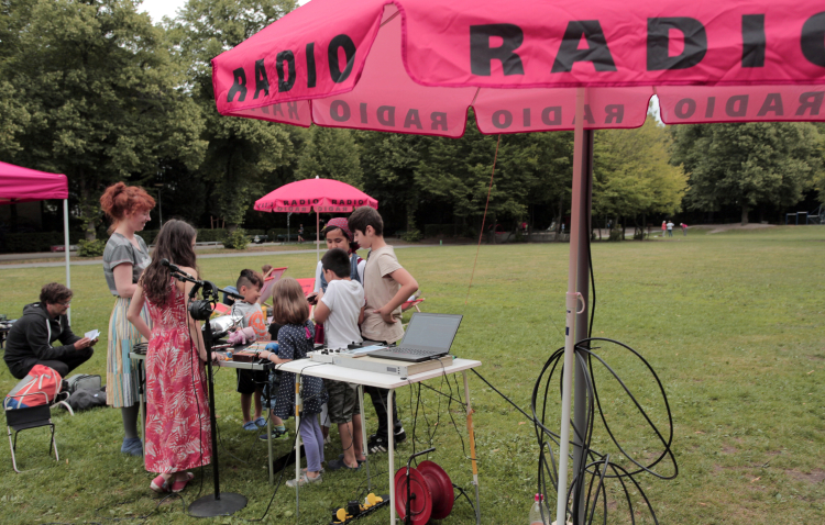 There are several tables with electronic radio equipment on a lawn. Several children are standing around the tables, interacting with them. Between them are magenta-colored parasols with the word "Radio" on them.