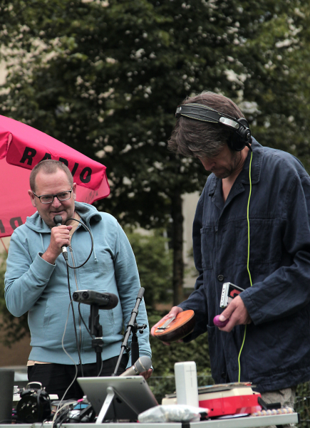 Ein Mann mit Köpfhörern steht an einem Tisch mit Mischpult und elektronischem Radio-Equipment. Neben ihm stehend der Künstler Ralf Homann mit einem Mikrofon.