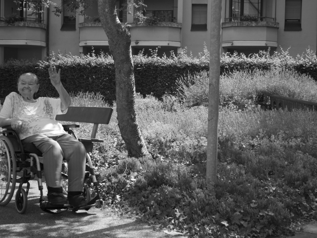 An old lady sitting in a wheelchair, her hand raised in a wave. In the background, plants and trees and the balconies of an apartment block.