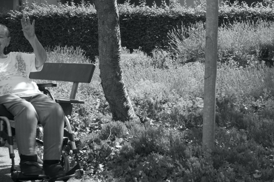 An old lady sitting in a wheelchair, her hand raised in a wave. In the background, plants and trees and the balconies of an apartment block.