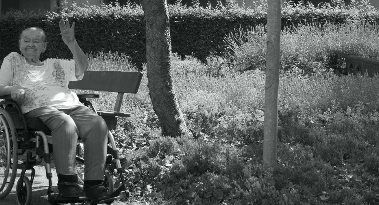 An old lady sitting in a wheelchair, her hand raised in a wave. In the background, plants and trees and the balconies of an apartment block.
