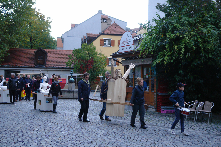 Procession with several people at Wiener Platz. Some participants carry two female figures as procession objects, one made of wood and one made of plaster, the so-called "Consumer Values Madonnas".