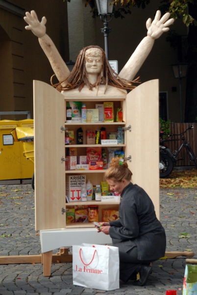 Image of the Konsum-Werte-Madonna (Consumer Values Madonna). A wooden figure with a body made from a cupboard with two doors and a head with long hair and outstretched arms. The body of the figure contains consumer products. The artist Stephanie Senge is sitting in front of it.