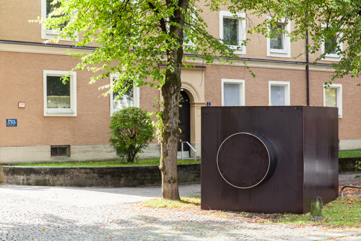 Auf einem Grünstreifen vor einem Apartmentblock ist eine Skulptur in Form eines dunklen, rechteckigen Baukörpers zu sehen, der an ein Kameragehäuse erinnert.