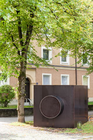 A sculpture in the form of a dark rectangular structure, reminiscent of a camera housing, can be seen on a green strip in front of a residential building.