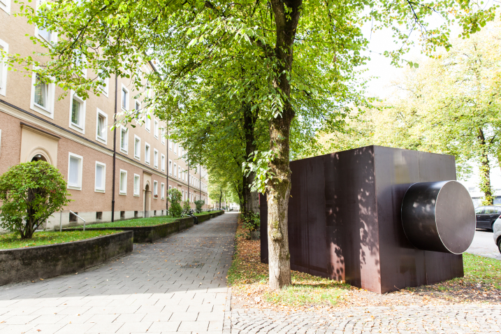 A sculpture in the form of a dark rectangular structure, reminiscent of a camera housing, can be seen on a green strip in front of a residential building.