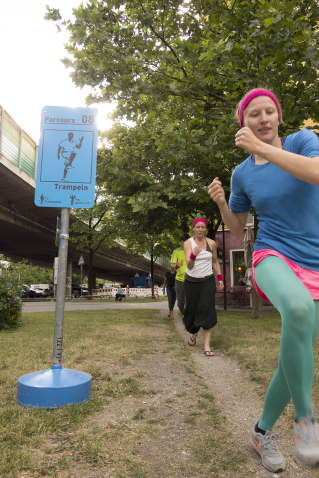 Four people in sportswear walk with firm steps in a row, next to a blue sign with the sporting instruction "Tramp". The Candidbrücke (Candid Bridge) can be seen in the background.