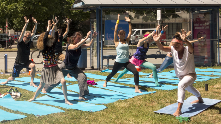 Eine Gruppe von Leuten in Yoga-Pose auf Yogamatten auf einer Wiese. Im Hintergrund eine Bushaltestelle und mehrspurige Straße.