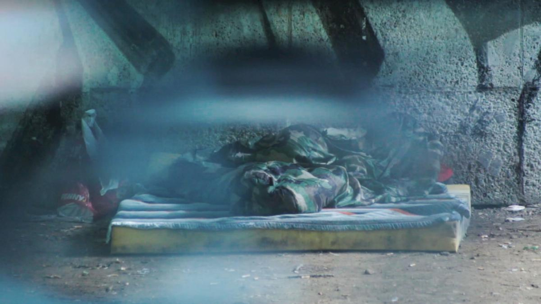 Screenshot of the video "Hors Champ". Blurry view of a homeless person, wrapped in a blanket, lying on a mattress outside in front of a wall, which appears to be a pillar of a bridge.