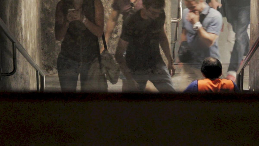 Screenshot of the video "Hors Champ". Blurred view into a stairwell of an underground railway staircase. Several people are climbing the stairs, a man in an orange-coloured top is sitting on the stairs.