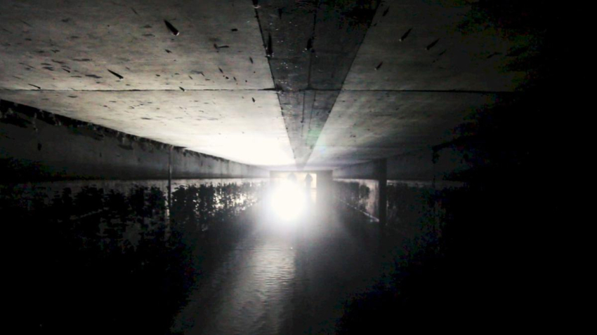 Screenshot of the video "Hors Champ". View through a dark shaft. Two people are standing on the opposite side, one of them is shining a torch.