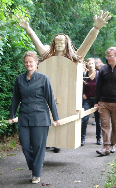 Prozession mit mehreren Menschen auf einem Spazierweg am Wald. Zwei Teilnehmer tragen als Prozessionsgegenstand eine kastenartige Frauenfigur aus Holz, die sogenannte "Konsum-Werte-Madonna".