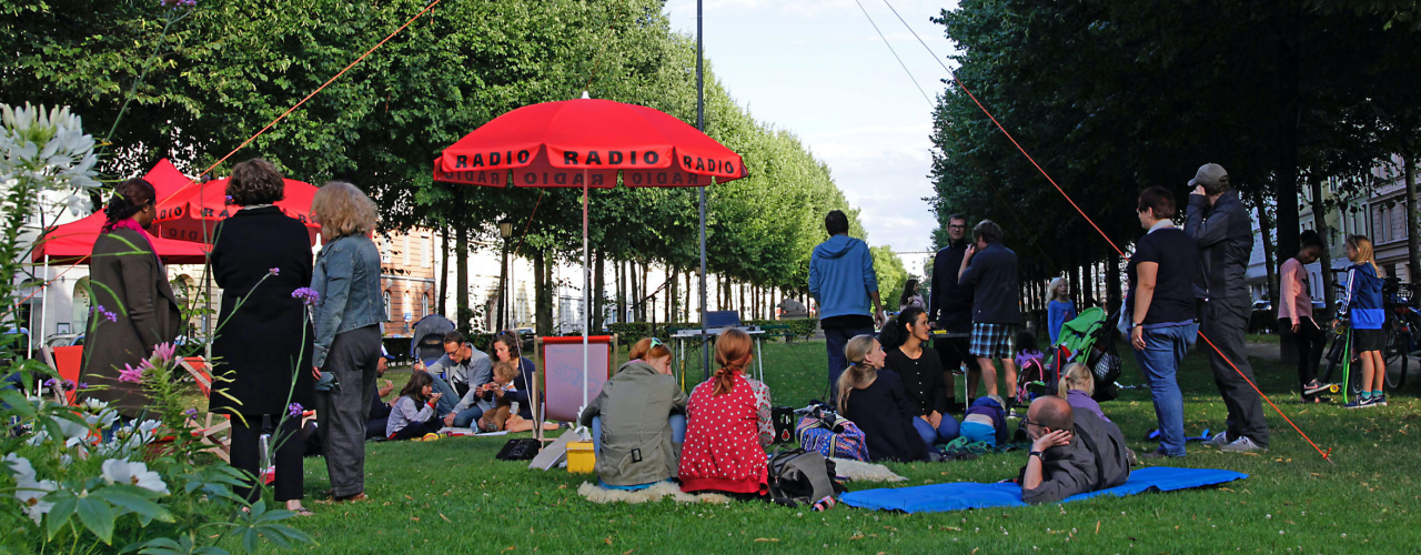 Lawn on Bordeauxplatz, with a radio antenna on a mast in the middle. Grouped around it are tables with radio equipment, parasols with the word "Radio" on them and deckchairs. People sit in the chairs, stand and sit on blankets.