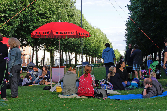 Lawn on Bordeauxplatz, with a radio antenna on a mast in the middle. Grouped around it are tables with radio equipment, parasols with the word "Radio" on them and deckchairs. People sit in the chairs, stand and sit on blankets.