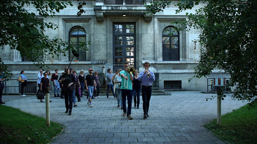 Eine Gruppe von Menschen folgt zwei Musikern mit Blasinstrumenten, die diesem Marsch vorangehen. Im Hintergrund zu sehen der Seiteneingang des Zentralinstituts für Kunstgeschichte in der Katharina-von-Bora-Straße 10.