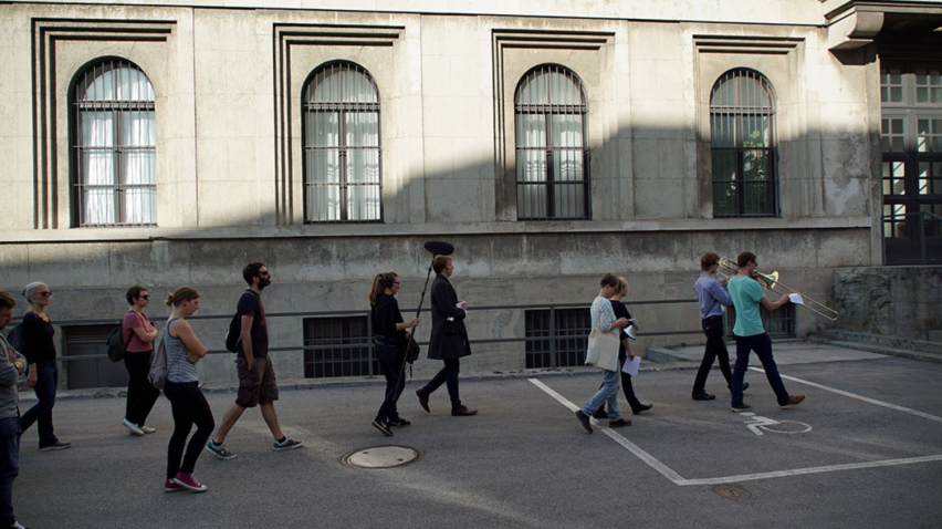 Eine Gruppe von Menschen folgt zwei Musikern mit Blasinstrumenten, die diesem Marsch vorangehen. Im Hintergrund zu sehen der Seiteneingang des Zentralinstituts für Kunstgeschichte in der Katharina-von-Bora-Straße 10.