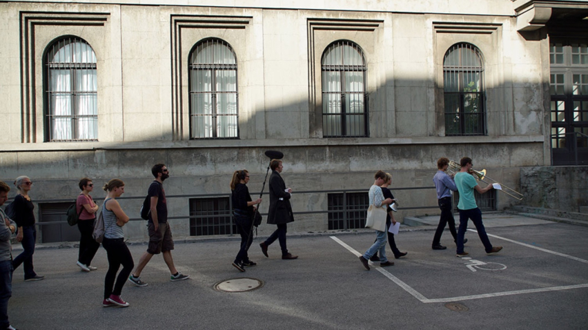 A group of people follow two musicians with wind instruments leading this march. The side entrance to the Zentralinstitut für Kunstgeschichte at Katharina-von-Bora-Straße 10 can be seen in the background.