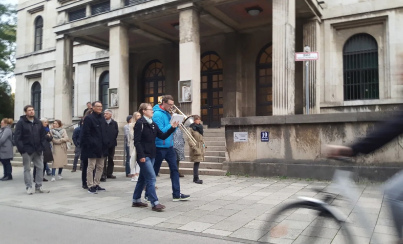 Eine Gruppe von Menschen folgt einem Musiker mit Blasinstrument, der diesem Marsch vorangeht. Im Hintergrund zu sehen der Haupteingang des Zentralinstituts für Kunstgeschichte in der Katharina-von-Bora-Straße 10.