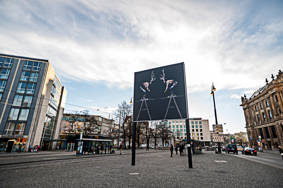 View of the billboard on Lenbachplatz looking towards the city centre with the motif "Homo Alpinus" by Stefano Giuriati. The motif shows on black background a mirror image of the artist Stefano Giuriati in a wrestling jersey with a deer antler on his head standing on a ladder. He is giving the impression that he is entering into a fight with himself.