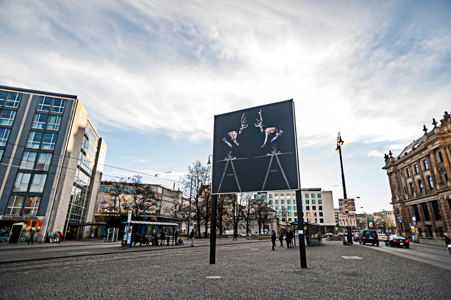 Ansicht des Billboards am Lenbachplatz Richtung stadteinwärts mit dem Motiv "Homo Alpinus" von Stefano Giuriati. Das Motiv auf schwarzem Hintergrund zeigt gespiegelt das gleiche Bild des Künstlers Stefano Giuriati in Ringertrikot mit einem Hirschgeweih auf dem Kopf auf einer Leiter stehend. Es wirkt, als würde dieser in einen Kampf mit sich selbst treten.