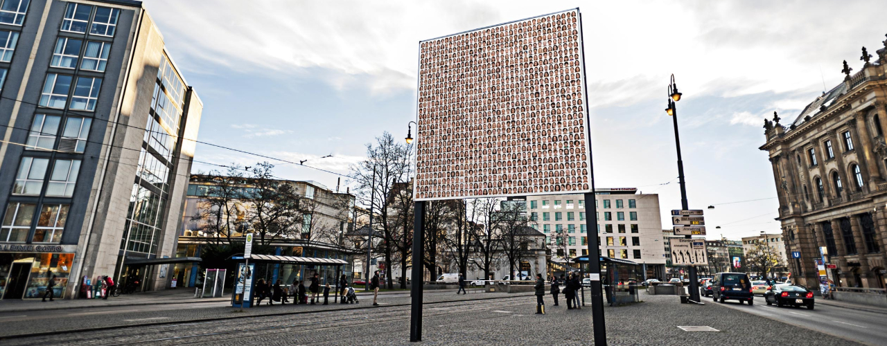 Side view of the billboard on Lenbachplatz with the motif "Israelisches Kollektivportrait" by Roland Fischer. The motif shows portraits of men and women arranged in rows, giving the impression of a collective portrait.