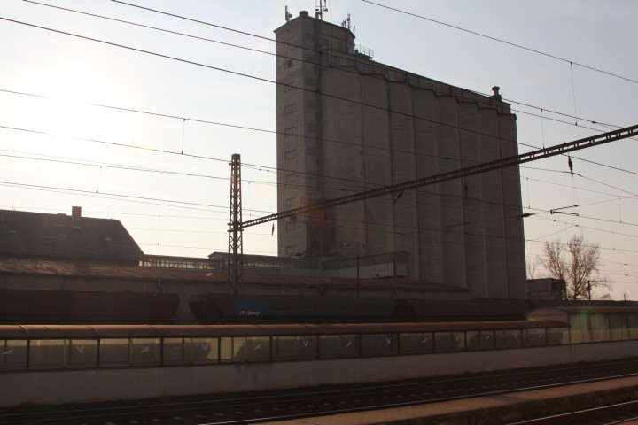 View from a train window onto the tracks and a tall industrial building in Bohusovice nad Ohri, Czech Republic.