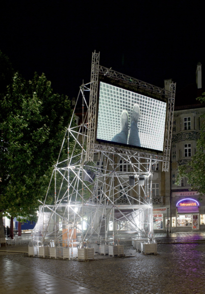 Munich's Rindermarkt at night. There is a metal scaffolding with a mounted video screen on which the video work "Top View" by Nevin Aladağ is shown.
