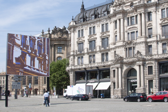 View of the billboard on Lenbachplatz looking towards the city with the motif "hide and seek" by Angela Stauber. The motif is a mirror image of the building to the right of the billboard. The architectural forms of the historic façade are simplified using the woodcut technique and abstracted into a block-like aesthetic of coloured surfaces in white, yellow and blue.