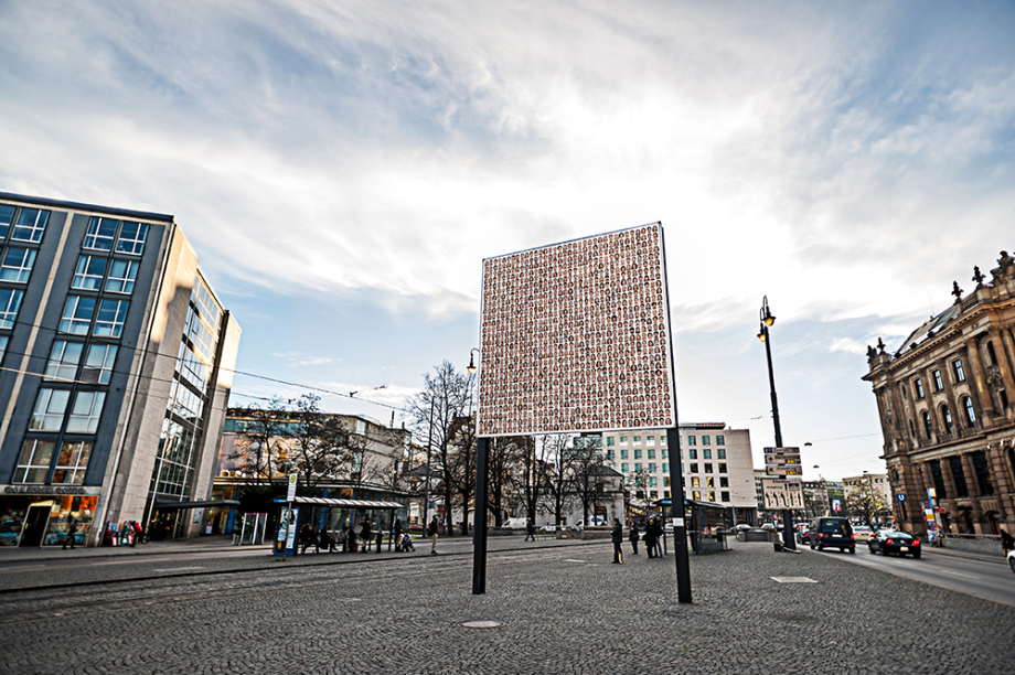 Side view of the billboard on Lenbachplatz with the motif "Israelisches Kollektivportrait" by Roland Fischer. The motif shows portraits of men and women arranged in rows, giving the impression of a collective portrait.