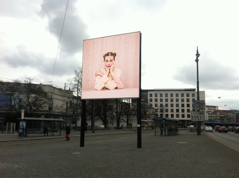 Diagonale Ansicht des Billboards am Lenbachplatz stadteinwärts blickend mit dem Motiv "Dreiarmige Frau" von Susanne Steinmaßl. Das Motiv zeigt eine blonde Frau in blassrosa Bluse vor einer rosanen gemusterten Tapete sitzend. Zwei Arme hat sie auf einen rosa Tisch vor sich abgestützt und stützt ihr Gesicht in beide Hände. Zudem hat die Frau einen dritten Arm, den sie auf den Tisch stütz und mit der Hand ihre Schulter umfängt.