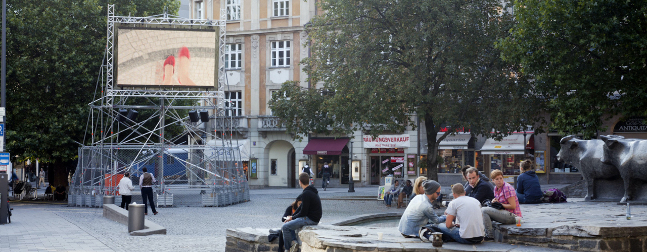 Auf dem Münchner Rindermarkt steht ein Metallgerüst mit einem aufmontierten Videoscreen, auf dem die Videoarbeit "Top View" von Nevin Aladağ gezeigt wird.