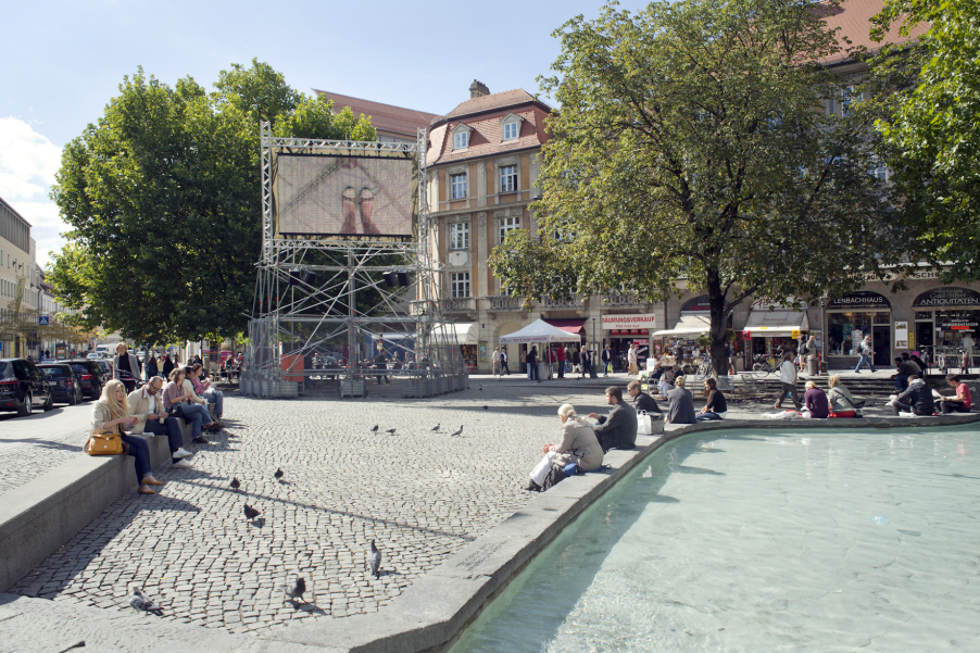 Auf dem Münchner Rindermarkt steht ein Metallgerüst mit einem aufmontierten Videoscreen, auf dem die Videoarbeit "Top View" von Nevin Aladağ gezeigt wird.