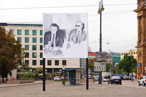 Das Motiv auf dem Billboard am Lenbachplatz zeigt die Tuschezeichnung einer Fotografie eines Treffens zwischen Franz Josef Strauß und Alexander Schalck-Golodkowski, Leiter des DDR-Außenhandelsministeriums.