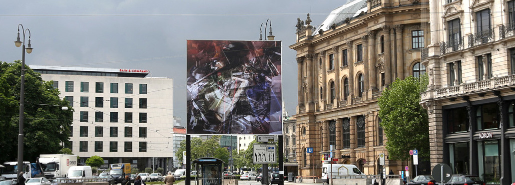 Frontale Ansicht des Billboards am Lenbachplatz stadteinwärts blickend. Das mehrfachbelichtete Foto auf dem Billboard zeigt eine Ansammlung von entsorgten Dinge in Containern. Bei längerem Hinsehen können so mehrere Schichten an unterschiedlichen Gegenständen, von Elektroschrott über Metallstangen bis hin zu Sitzpolstern, ausgemacht werden.