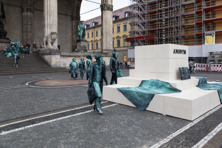 Fotografie eines Kunstprojektes von Silke Wagner am Odeonsplatz vor der Feldherrnhalle. Die Installation ist ein lebendes Denkmal, das die Illusion einer Skulptur erzeugen soll: In der Mitte des Odeonsplatzes stehen pyramidenartig gestapelte weiße Sockel. Auf den unteren zwei Sockeln liegen zwei Decken. Zudem steht dort eine Gedenktafel mit der Aufschrift "Zum Gedenken an die Opfer von sexueller Gewalt im Krieg" und auf dem höchsten Sockelblock ist in Großbuchstaben das Wort "Anonym" angebracht, was gleichzeitig auch der Titel des Kunstprojektes ist. Von den Treppen der Feldherrnhalle steigen eine Reihe von Performer*innen, die in Richtung der Sockel laufen. Sie sind in grün-graue Kostüme gekleidet, die sich der Grünspan-Bronzepatina der Feldherrenskulpturen in der Feldherrnhalle anpassen.