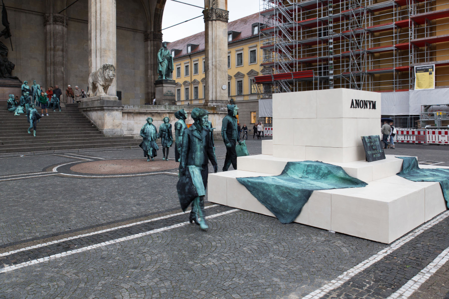 Photograph of an art project by Silke Wagner on Odeonsplatz in front of the Feldherrnhalle. The installation is a living monument intended to create the illusion of a sculpture: In the centre of Odeonsplatz are white plinths stacked like pyramids. Two blankets lie on the two lower plinths. There is also a memorial plaque with the inscription "In memory of the victims of sexual violence during the war" and the word "Anonymous" in capital letters on the highest block of plinths, which is also the title of the art project. From the steps of the Feldherrnhalle, a series of performers walk towards the plinths. They are dressed in green-grey costumes that match the verdigris bronze patina of the Feldherrnhalle sculptures.